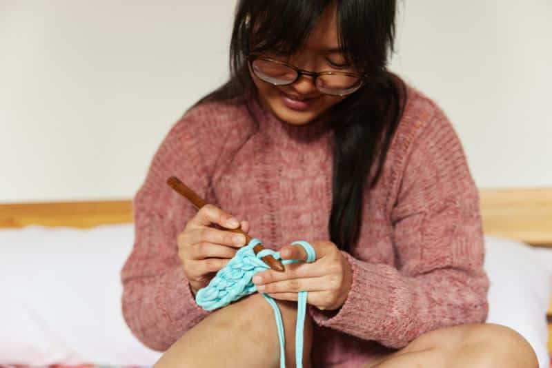 young woman in pink sweater sitting on bed crocheting